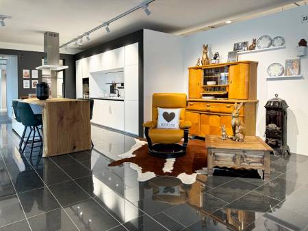 Offener Küchenraum in Weiß und Grau mit einer Insel aus Naturholz, einem gelben Ledersessel und einer klassischen Vitrine, kombiniert mit traditionellen Wohnaccessoires auf glänzendem dunklen Fliesenboden.